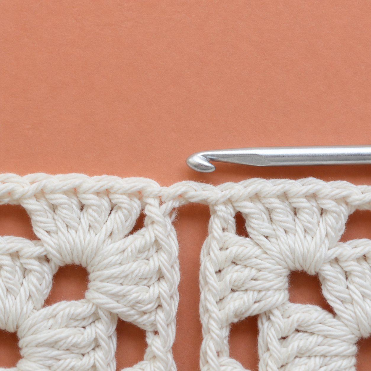 AI-assisted close-up showing a crochet hook aimed at the smaller open side chain space between granny-square clusters.